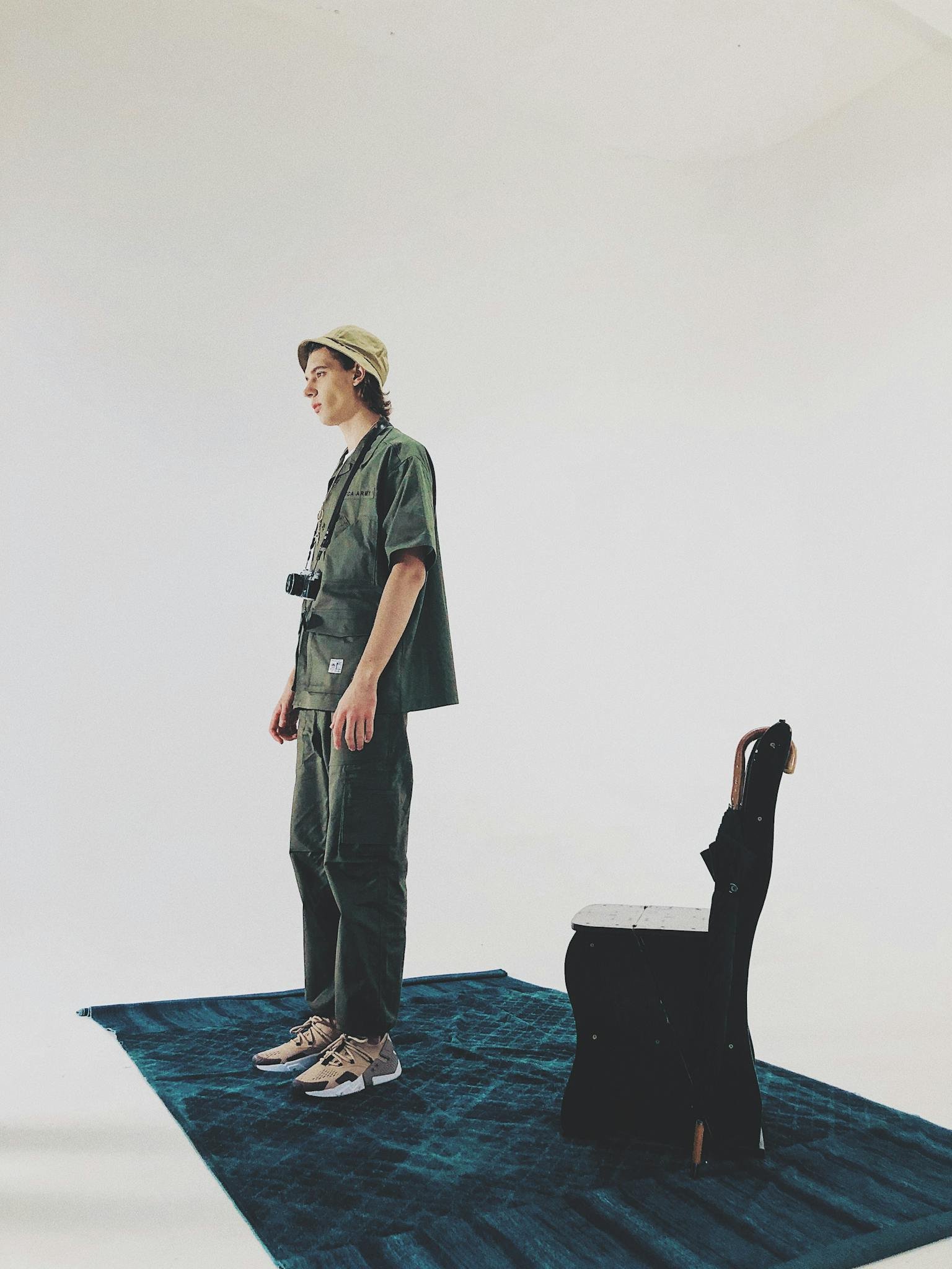 Man in stylish outfit standing beside a chair on a blue rug in a studio setting.